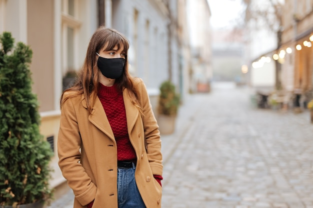 Beautiful young brunette dressed in casual clothing posing at city street in medical face mask. Concept of flu and coronavirus infection.