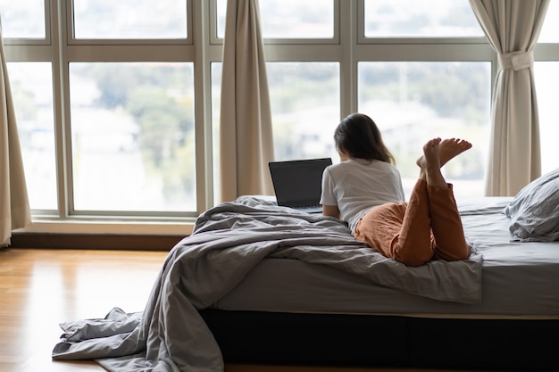 A beautiful young brunette girl is working on the laptop while lying on a bed by a panoramic window with a beautiful view from a high floor. Stylish modern interior. A cozy workplace.