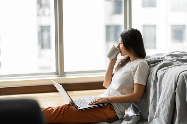 Beautiful young brunette girl working on a laptop and drinking coffee, sitting on the floor near the bed by the panoramic window