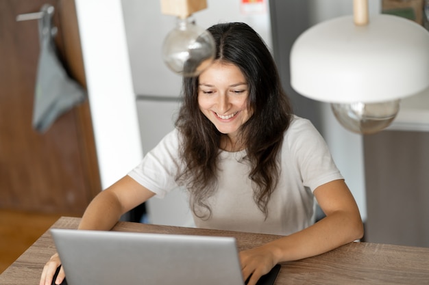 Beautiful young brunette girl working on laptop at home in the kitchen.