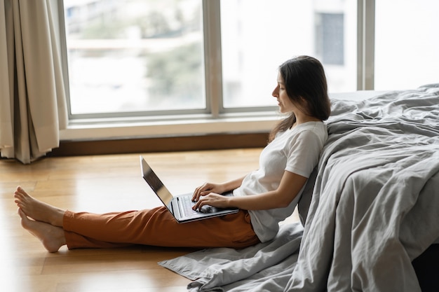 Beautiful young brunette girl working on a laptop, sitting on the floor near the bed by the panoramic window with a beautiful view from the high floor. A cozy workplace. Shopping on the Internet.