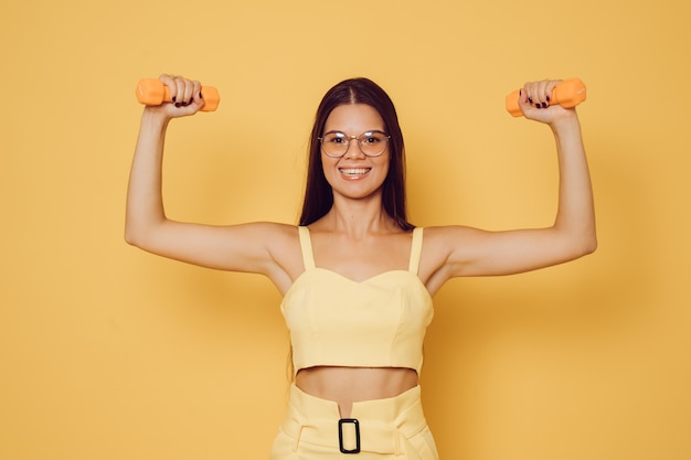 Beautiful young brunette in glasses dressed in yellow top and trousers, broad smiling, lifted dumbbells shows off her biceps happy to exercise and be healthy. Confident people concept.