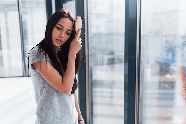 Beautiful young brunette in grey shirt standing near window and posing for a camera.