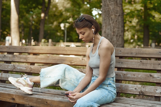 ベンチの公園で音楽を聴いている美しい若いブルネットの流行に敏感な女性。