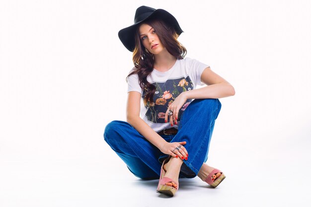 Beautiful young brunette in jeans bell bottom white t-shirt and black hat ,sitting on floor