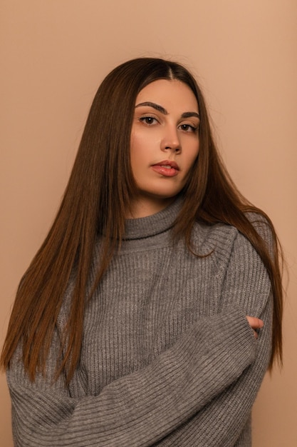 Beautiful young brunette model girl in a gray fashionable knitted sweater on a beige background. Girl in outerwear in the studio