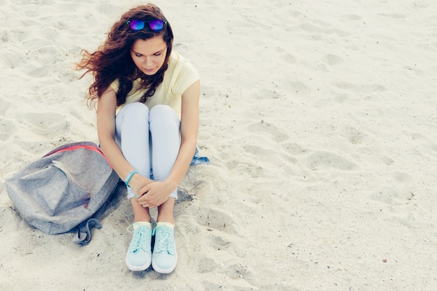 Bella bruna giovane in occhiali da sole, t-shirt e jeans seduti sulla spiaggia