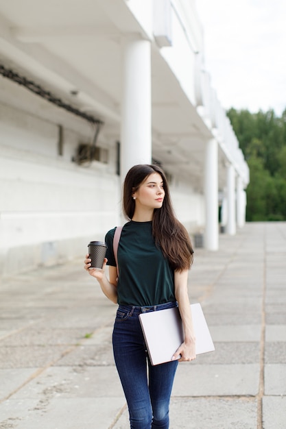 Beautiful young brunette woman carrying backpack and drinking takeaway coffee while walking outdoors