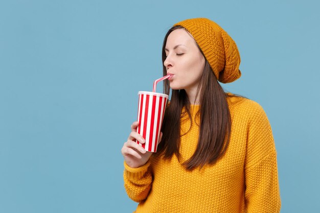 Beautiful young brunette woman girl in yellow sweater hat\
posing isolated on blue background studio portrait. people sincere\
emotions lifestyle concept. mock up copy space. hold cup of soda or\
cola.