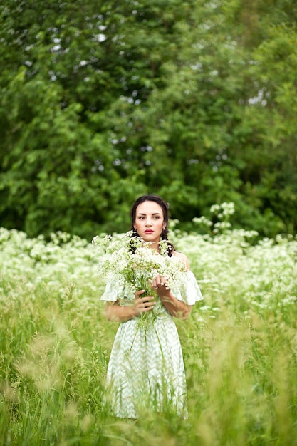 Beautiful young brunette woman in a green dress on the meadow with white flowers in a hand on a warm summer day