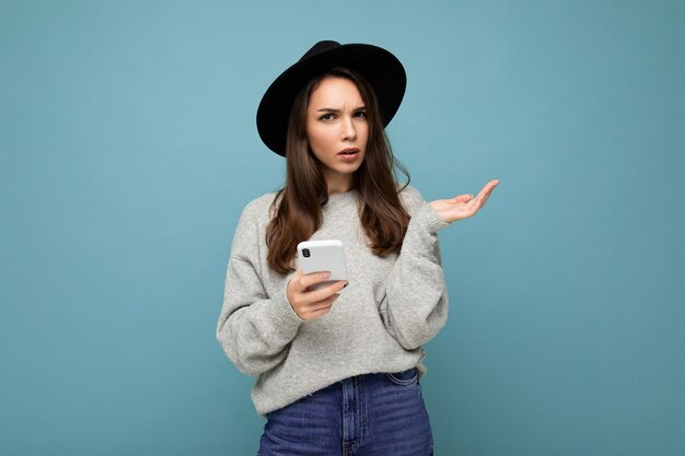 Beautiful young brunette woman thinking wearing black hat and grey sweater holding smartphone