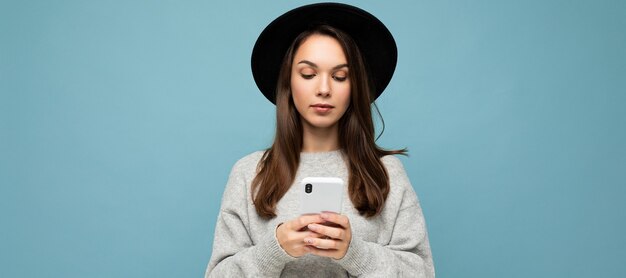 スマートフォンを保持している黒い帽子と灰色のセーターを着ている美しい若いブルネットの女性