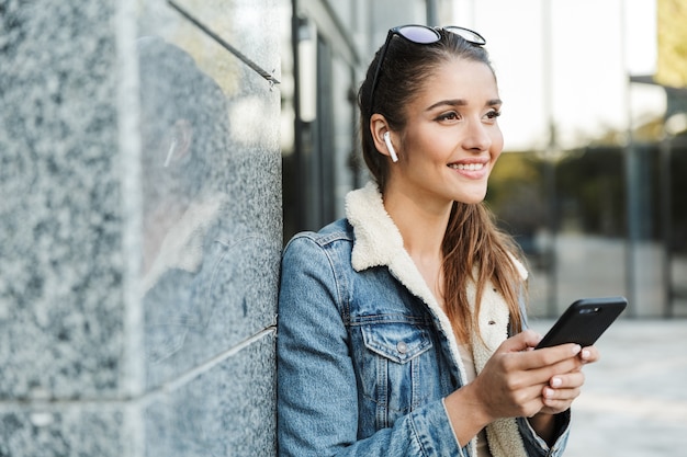 Beautiful young brunette woman wearing jacket, carrying backpack walking outdoors, using mobile phone