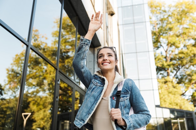 Beautiful young brunette woman wearing jacket, carrying backpack walking outdoors, waving hand