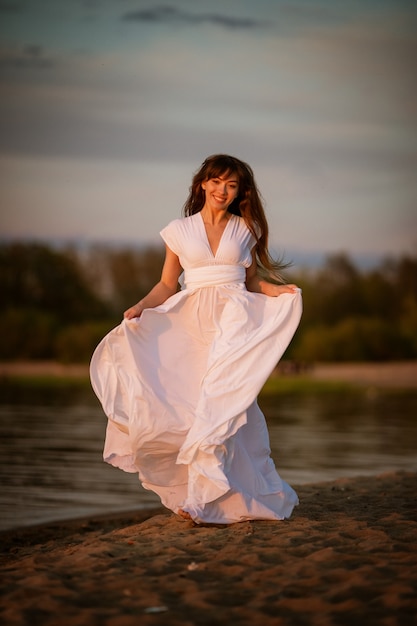 Bella giovane donna castana in un vestito bianco fluente sulla spiaggia sabbiosa la sera ritratto a figura intera in luce naturale