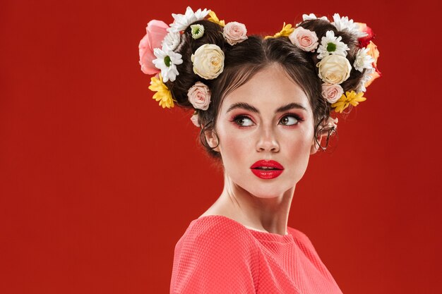 Beautiful young brunette woman with an amazing floral hairstyle wearing makeup posing isolated over red wall