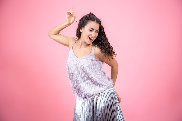 Beautiful young brunette woman with curly hair in a festive outfit on a pink wall  dancing with a smile on her face