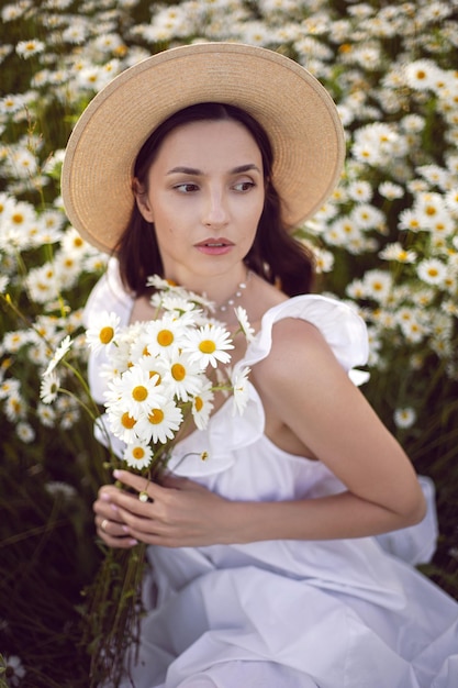 Beautiful young brunette woman with long hair in a hat and a white dress is holding a bouquet in her hands standing on a chamomile field at sunset in summer