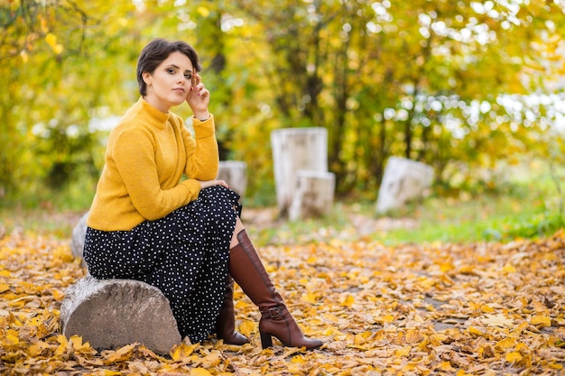 Bella giovane donna bruna in una gonna maglione lavorato a maglia giallo e stivali in posa nel parco autunnale e sorridente