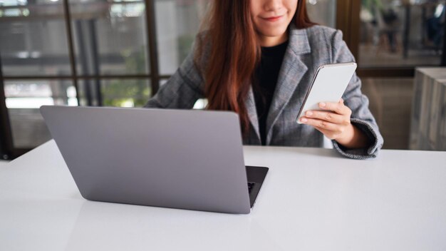 사무실에서 휴대전화와 노트북 컴퓨터를 들고 사용하는 아름다운 젊은 비즈니스 여성