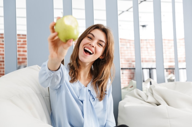 Beautiful young business woman sitting on a couch at the office, showing green apple