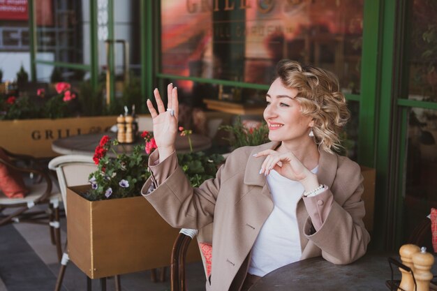 Beautiful young business woman sitting in a street cafe and waving her hand, greeting someone