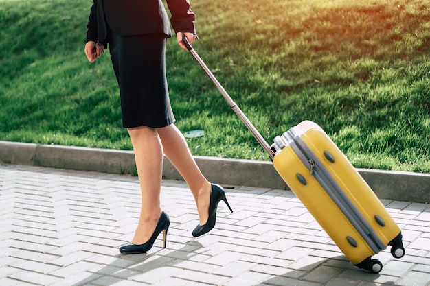 Beautiful young business woman in a suit and in shoes walking on a city street with a yellow travel suitcase