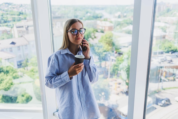 Красивая молодая деловая женщина разговаривает по смартфону и работает над новым проектом повышения производительности для компании, проводя свободное время в коворкинг-центре, наслаждаясь видом из окна