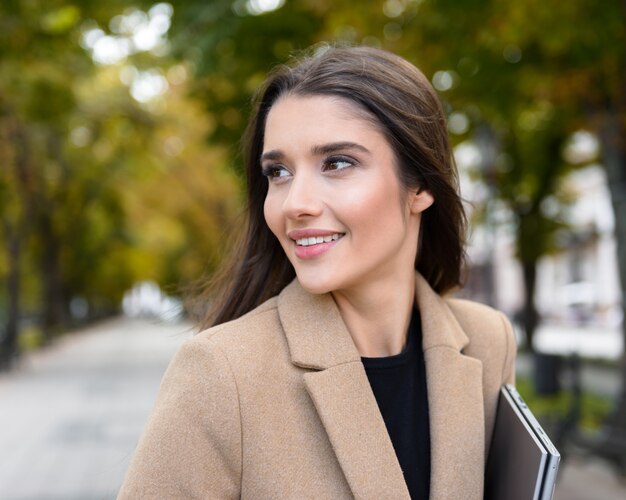 Красивая молодая деловая женщина в осеннем пальто гуляет на открытом воздухе в парке, неся ноутбук