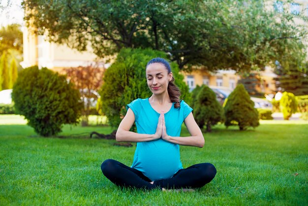 Beautiful young calm pregnant woman is sitting in the lotus position and meditating while practicing yoga in the park