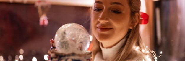 Beautiful young candid woman holding in hands near face and admire snow globe or dome glass ball and enjoyment an inspiring mood relaxing at home on merry christmas or happy new year eve night banner