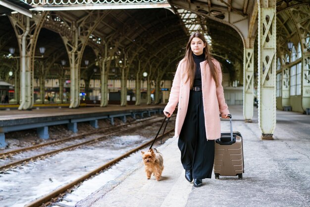 駅旅行で電車を待っている犬とスーツケースを持つ美しい若いカジュアルな観光客の女性...