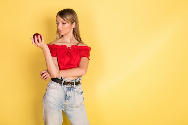 Beautiful young caucasian beauty blondie girl portrait with apple on yellow wall