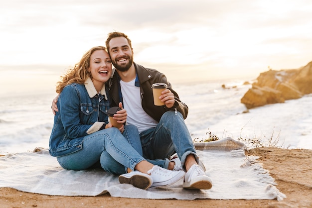 beautiful young caucasian couple drinking coffee together while walking by seaside