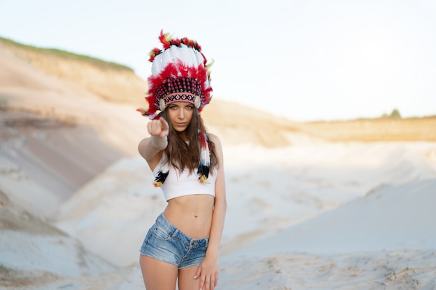 A beautiful young Caucasian girl in a white top and denim shorts. On her head  wearing an Indian hat Roach. Standing in the desert. 