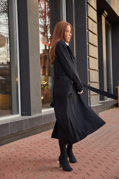 Beautiful young caucasian red headed girl in stylish black coat walking outdoors