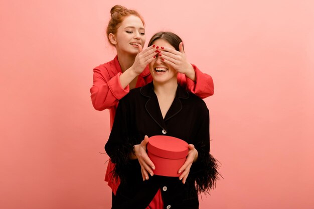 Photo beautiful young caucasian redhead girl closes eyes brunette friend holding gift on pink background concept of celebration happiness and emotions