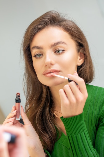 Beautiful young caucasian woman applying gloss to the lips looking in the mirror