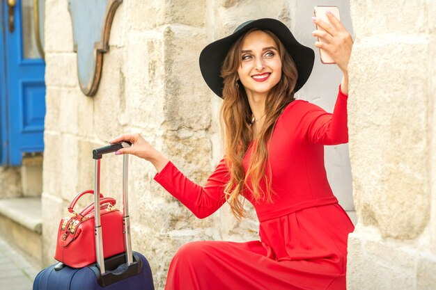 Красивая молодая кавказская женщина в черной шляпе, глядя на смартфон, улыбаясь и сидя на лестнице у двери на открытом воздухе.