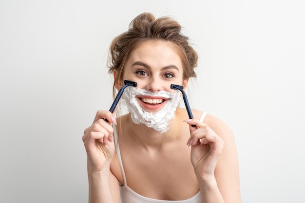 Beautiful young caucasian woman shaving her face by razor on white background. Pretty smiling woman with shaving foam and razor on her face.