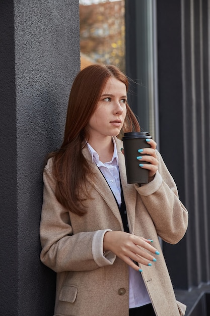 Beautiful young caucasian woman in stylish beige coat drinking coffee outdoors