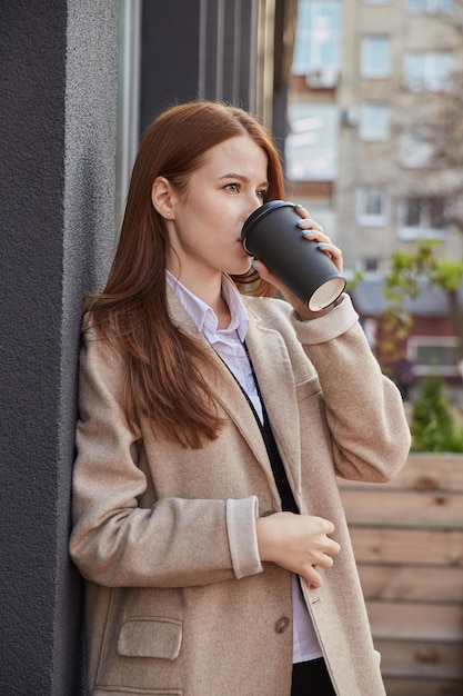 Beautiful young caucasian woman in stylish coat with long hair\
standing outdoors drinking coffee