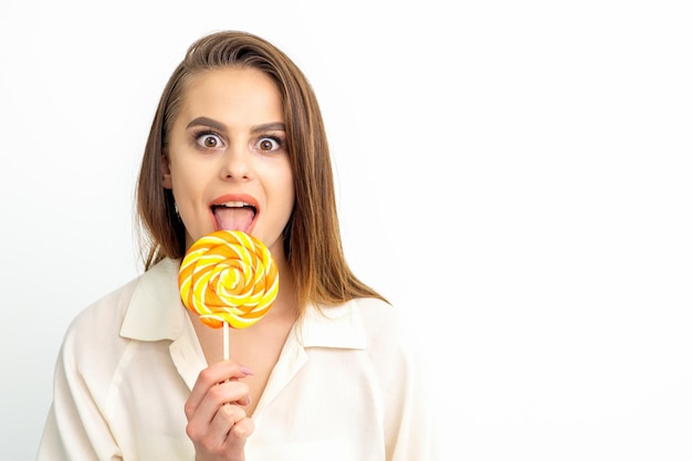 Beautiful young caucasian woman wearing a white shirt licking a lollipop on a white background