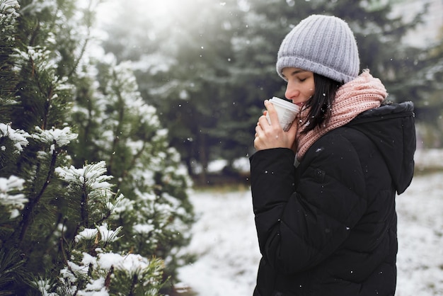 冬のジャケットを着た美しい若い白人女性は、雪の降る冬の日に公園や森の屋外でコーヒーやお茶を持ってビーニーグレーの帽子とピンクのスカーフを編んだ冬の気分