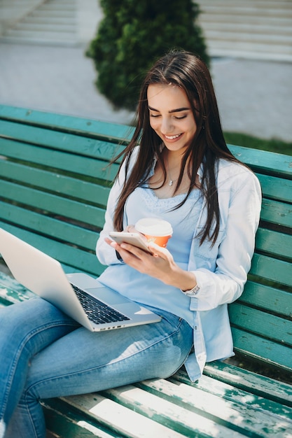 Bella giovane donna caucasica con lunghi capelli scuri che si siede su una panchina fuori a leggere su uno smartphone mentre si tiene un computer portatile sulle gambe sorridendo.