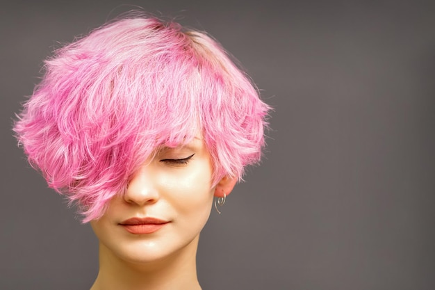 Beautiful young caucasian woman with short curly bob hairstyle dyed in pink color with closed eyes against dark gray background with copy space