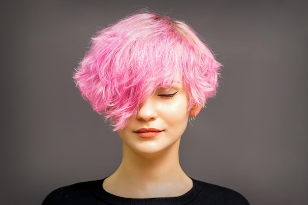 Beautiful young caucasian woman with short curly bob hairstyle dyed in pink color with closed eyes against dark gray background with copy space