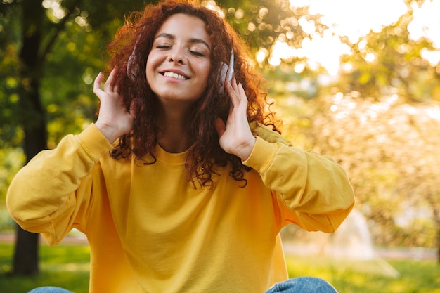 Bella giovane donna allegra che trascorre del tempo al parco, seduta su una panchina, ascoltando musica con le cuffie