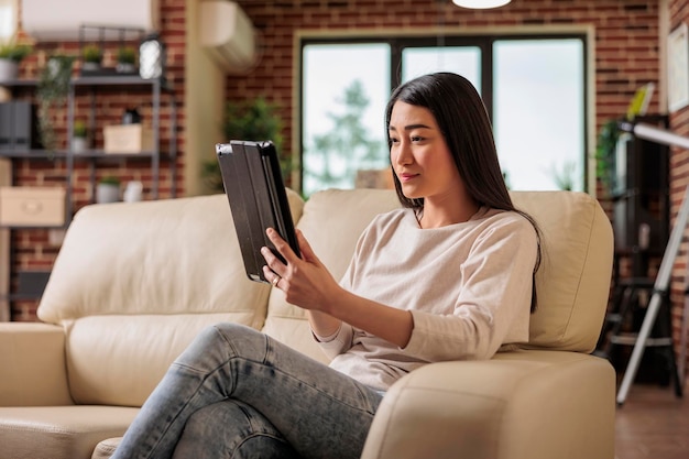 Beautiful young chinese woman using digital tablet at home, connecting internet web 3.0 smiling while sitting on couch entertainment device, browsing internet wireless device computer tablet using