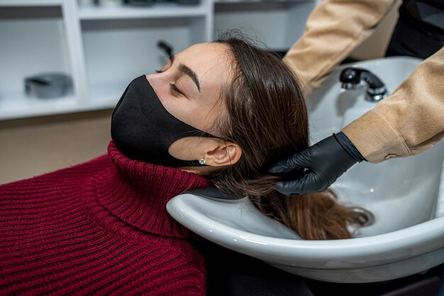 Foto una bellissima giovane cliente è venuta dal barbiere con una maschera speciale e il maestro prepara i capelli per il lavaggio durante l'epidemia concetto di parrucchiere